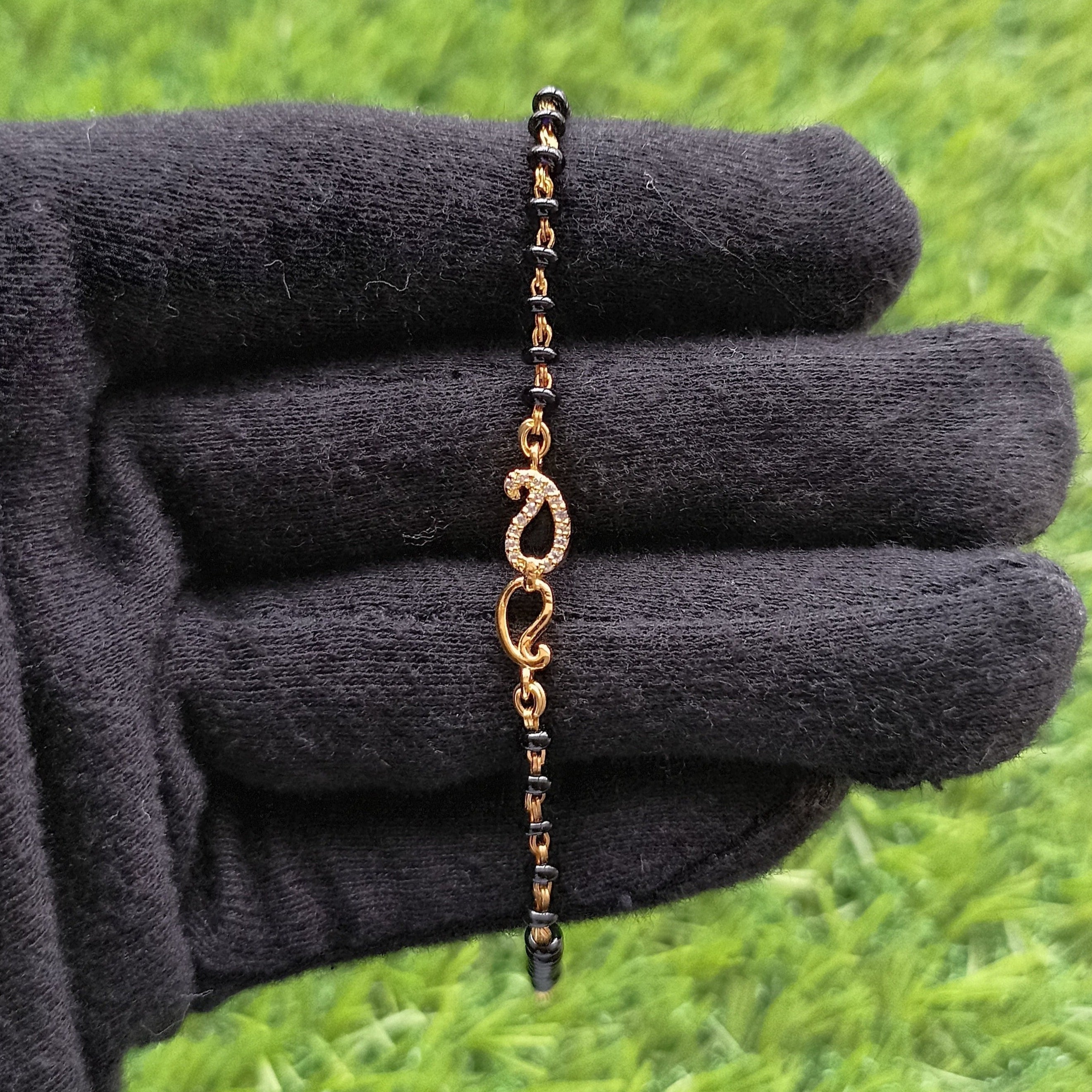 A hand mangalsutra bracelet with black beads and gold-colored leaf-shaped embellishments, displayed on a black glove against a grass like surface